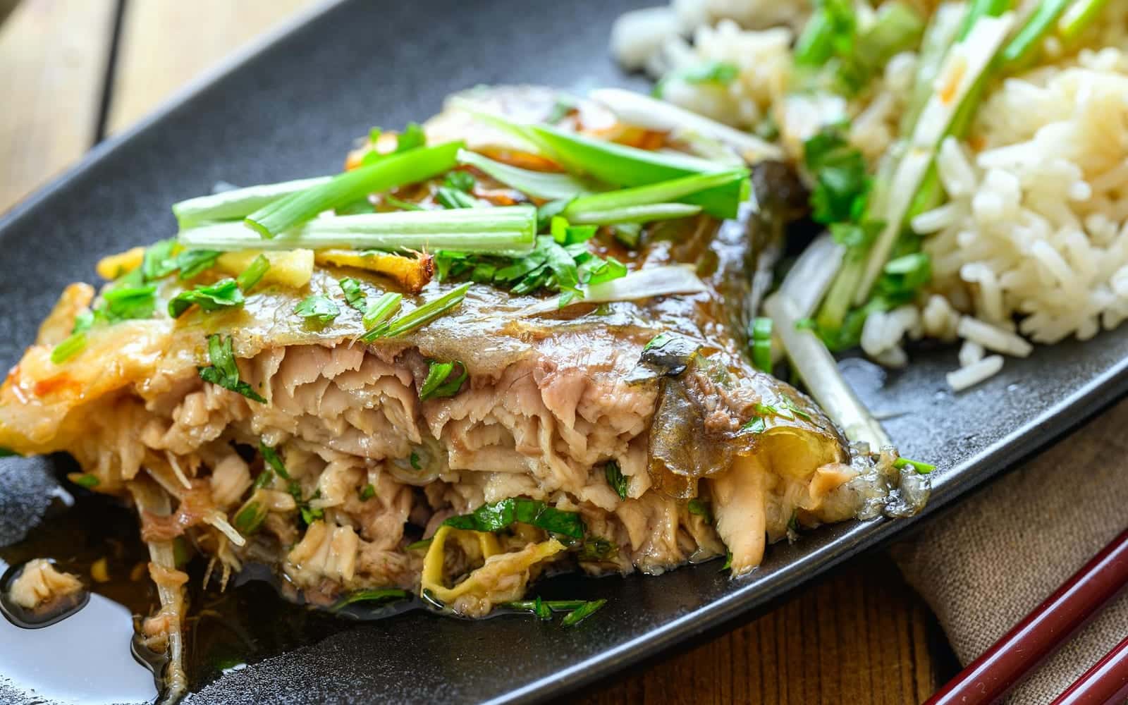 Chinese steamed whole fish with scallions, ginger, sauce, and rice on a black plate