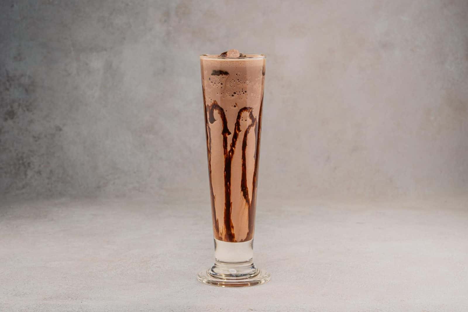 Chocolate smoothie in a tall glass with chocolate drizzle on a neutral background