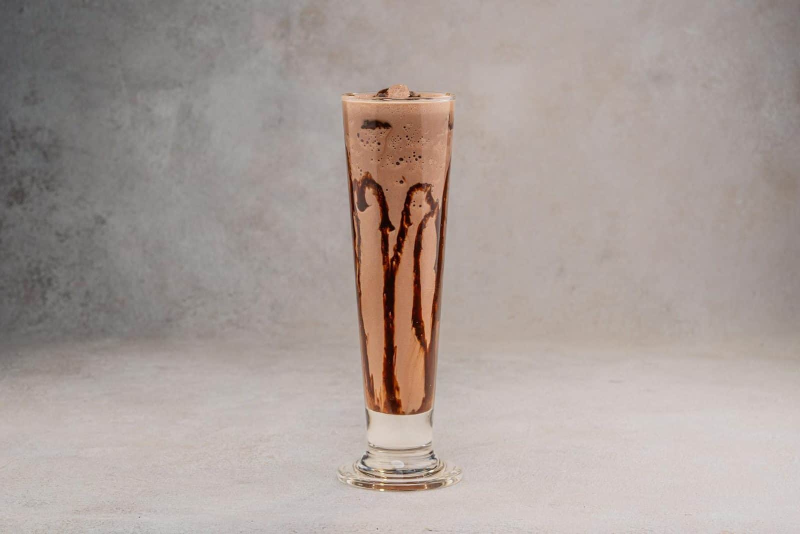 Chocolate smoothie in a tall glass with chocolate drizzle on a neutral background