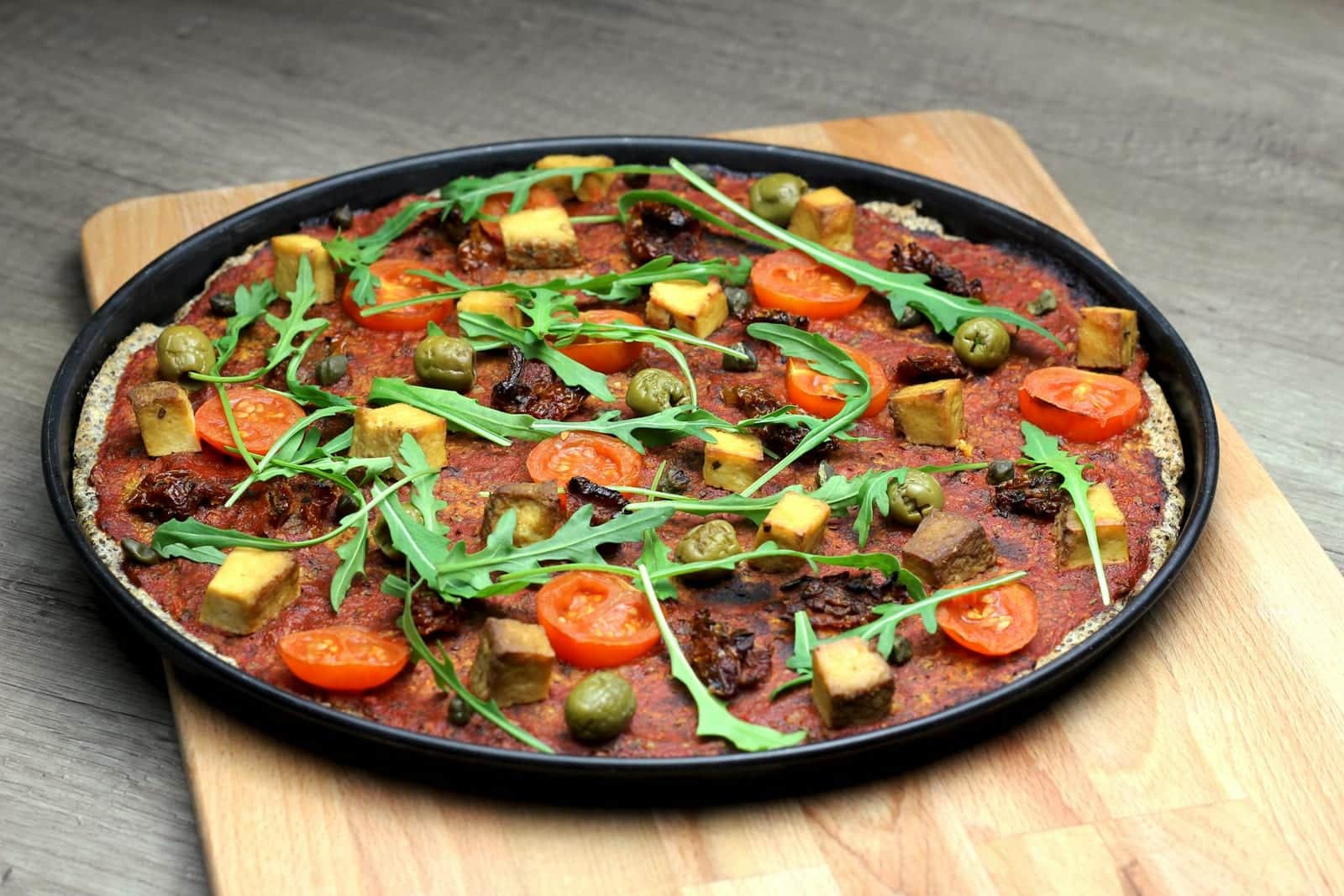 Vegan pizza topped with tomatoes, greens, olives, tofu, and vegetables on a dark pan