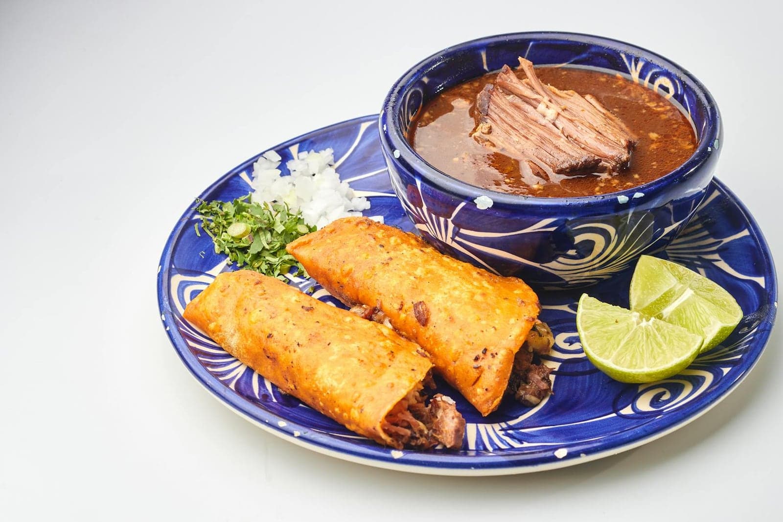 Birria tacos served with consomme, lime, onions, and cilantro on a blue plate