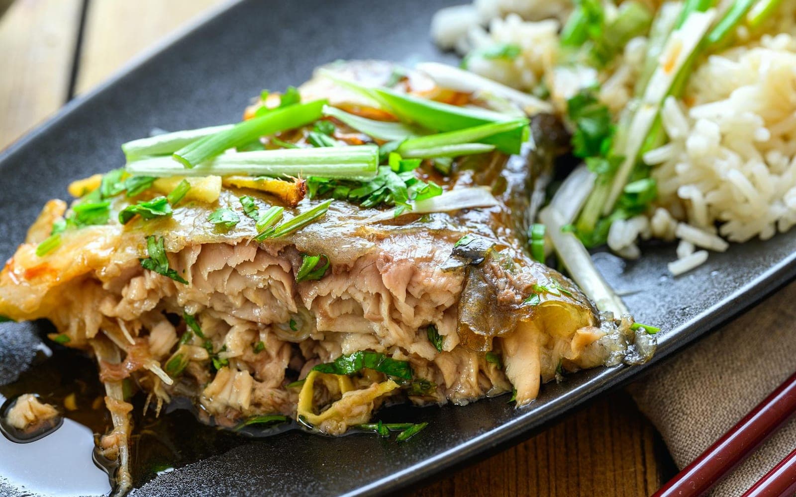 Chinese steamed whole fish with scallions, ginger, sauce, and rice on a black plate