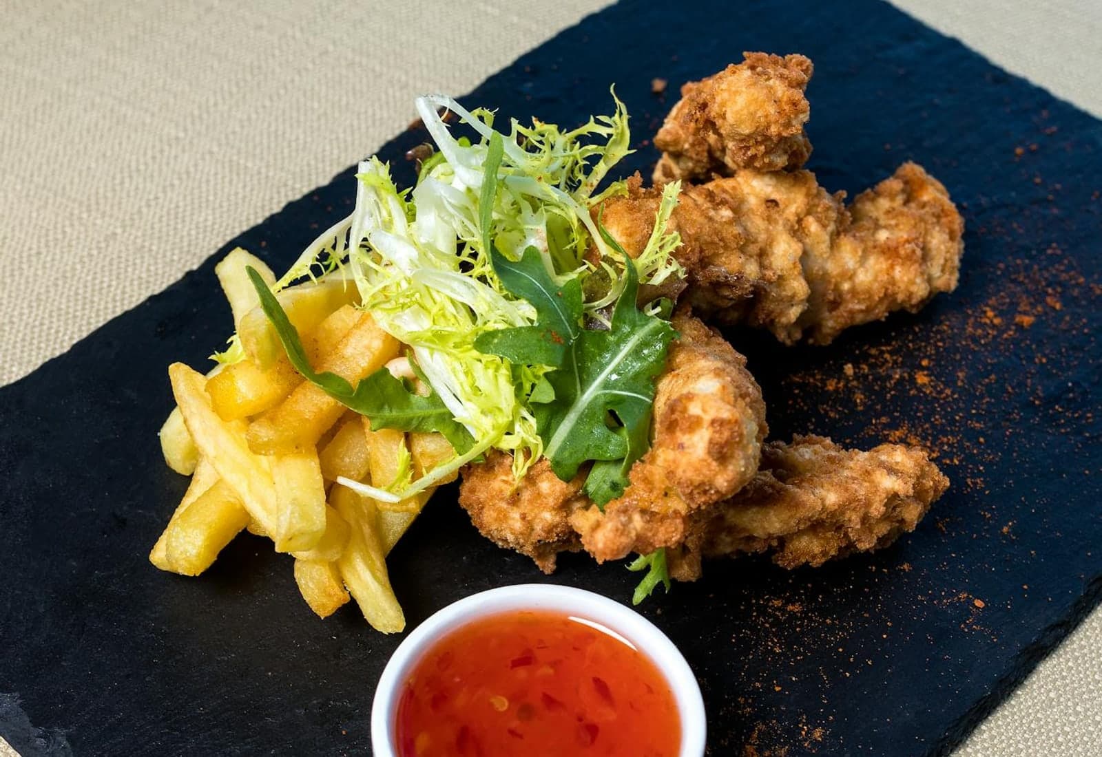 Crispy fried chicken pieces with dipping sauce, fries, and a small salad on a black plate