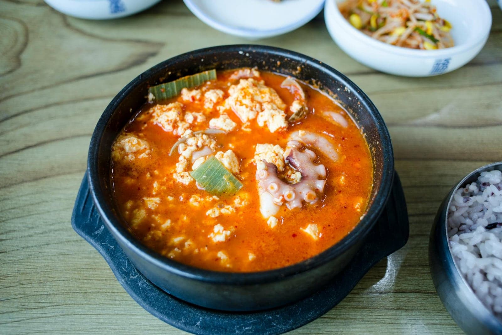 Sundubu jjigae Korean tofu stew in a black stone bowl with egg and side dishes