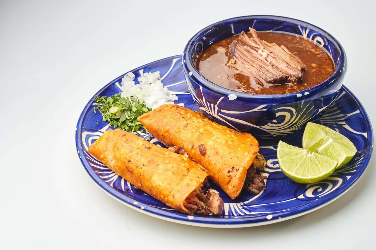 Birria tacos served with consomme, lime, onions, and cilantro on a blue plate