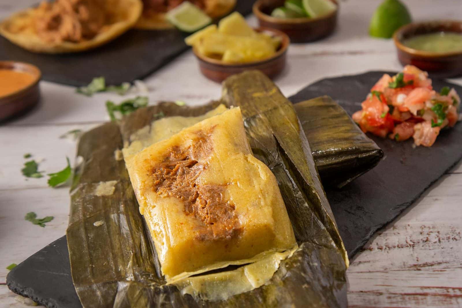 Chicken tamales verdes in corn husks with salsa and garnishes on a rustic table