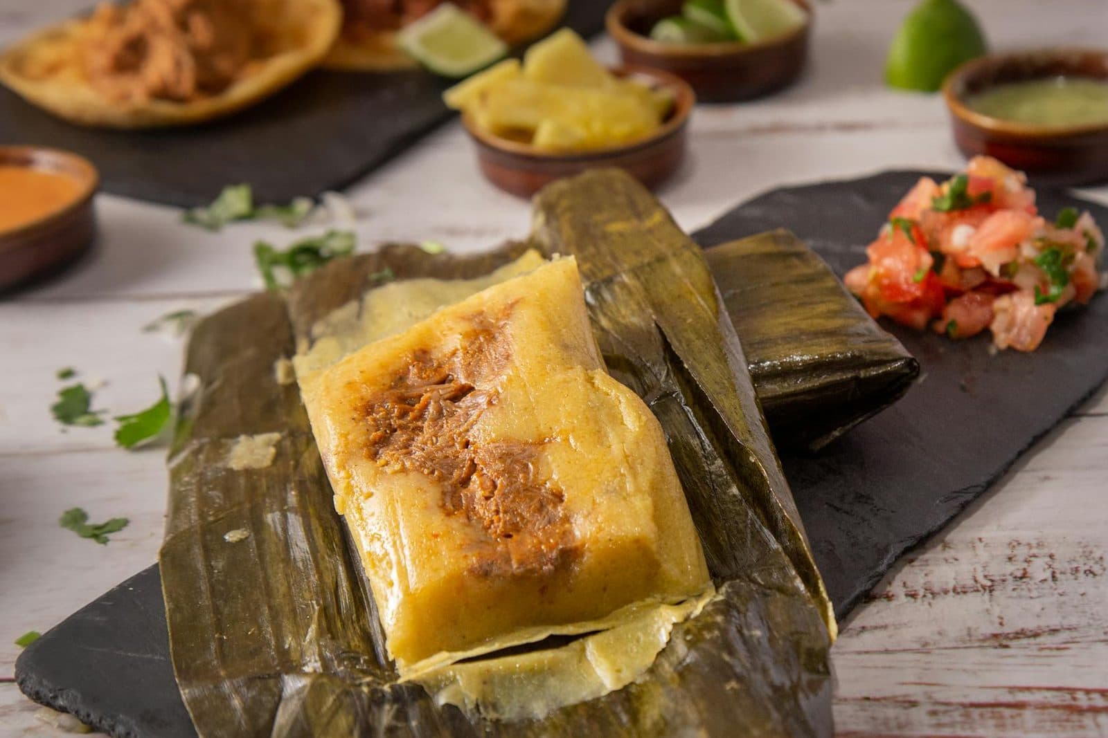 Chicken tamales verdes in corn husks with salsa and garnishes on a rustic table