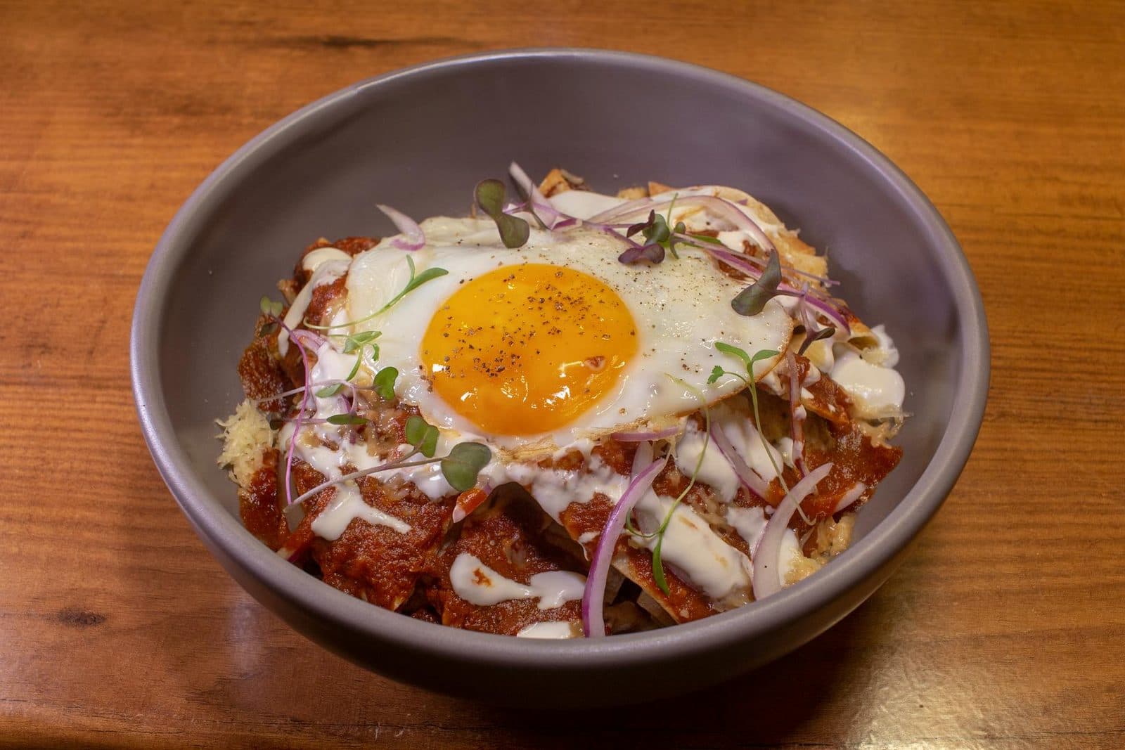 Chilaquiles rojos in a bowl with red salsa, fried egg, onion, cheese, and microgreens