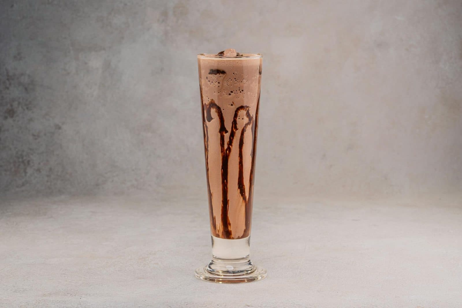 Chocolate smoothie in a tall glass with chocolate drizzle on a neutral background