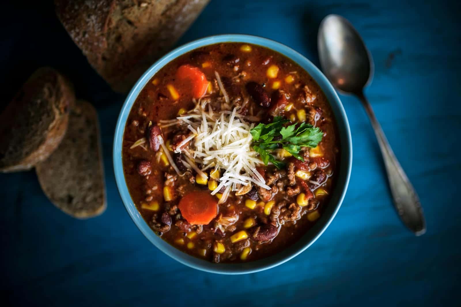 Classic beef chili in a blue bowl with beans, tomatoes, cheese, herbs, and bread on the side