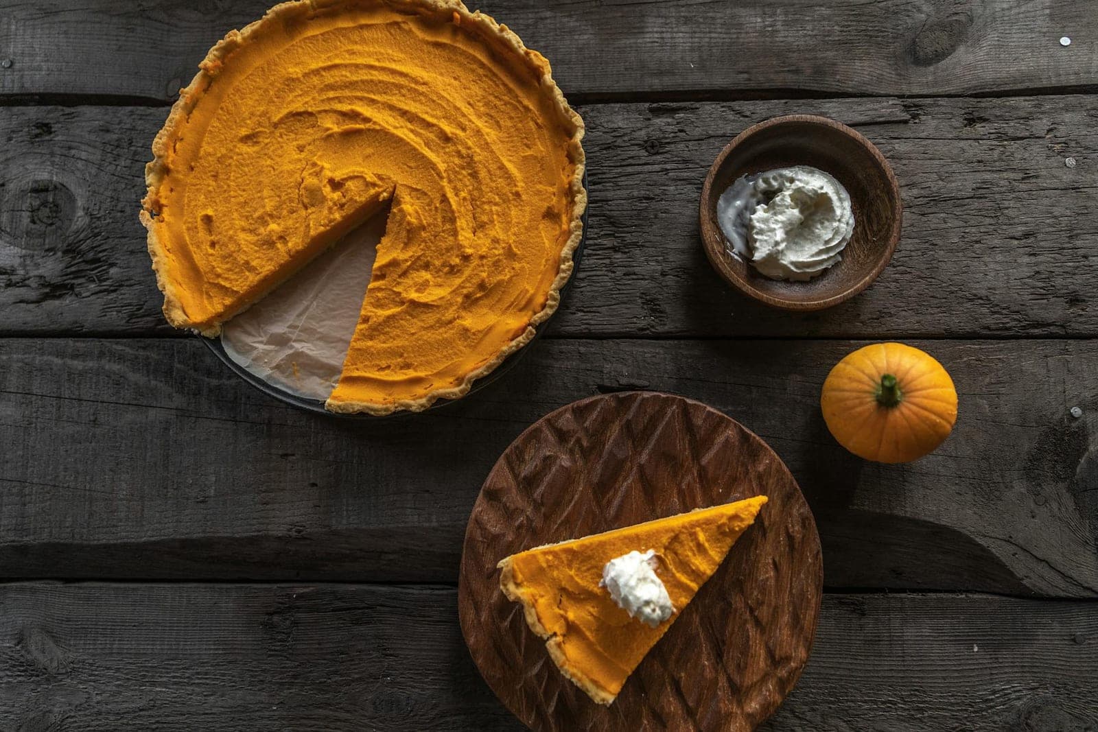 Classic pumpkin pie with a sliced wedge, whipped cream, and a small pumpkin on a rustic wood table
