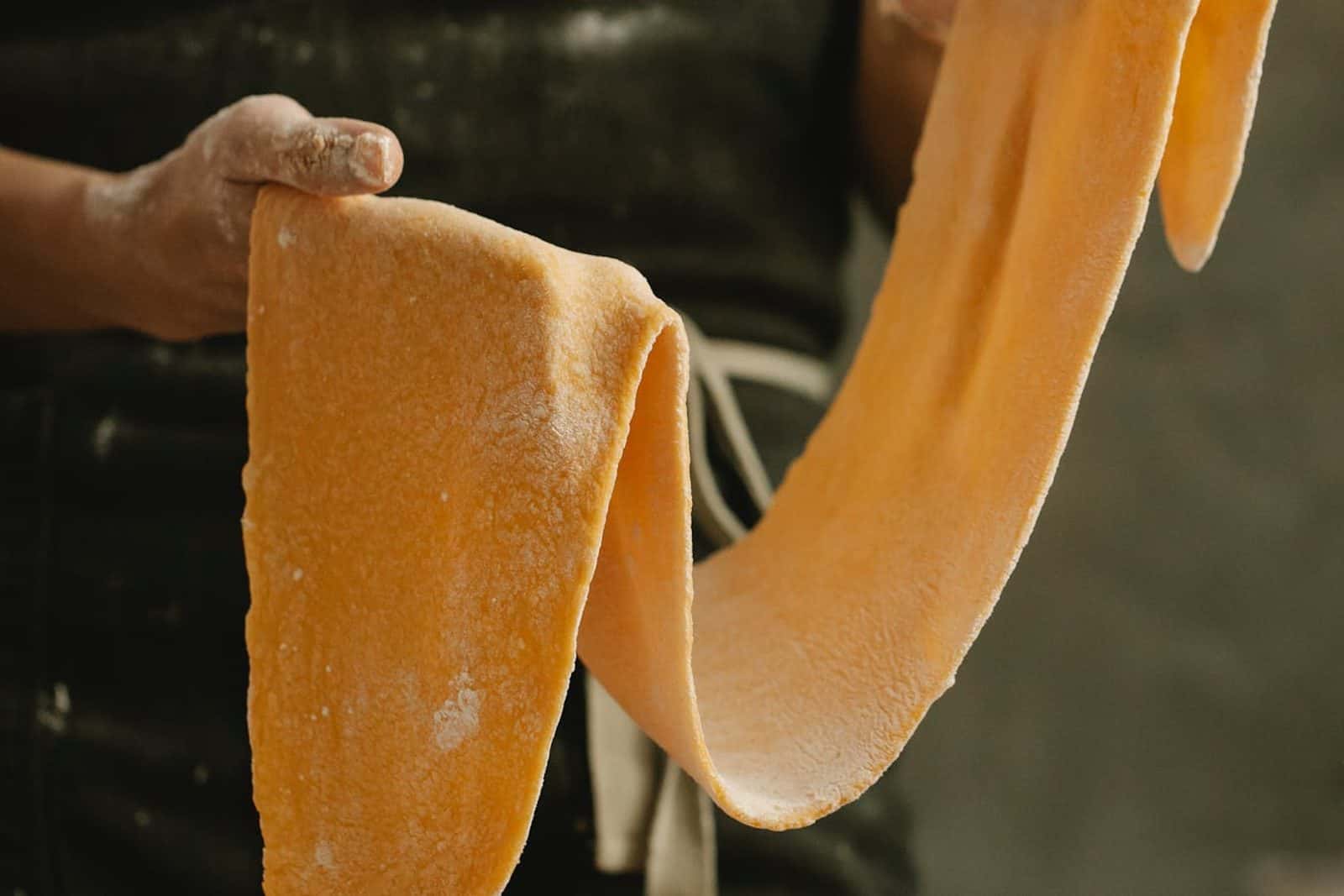 Fresh pasta dough sheet being stretched by hand with flour on a gray work surface