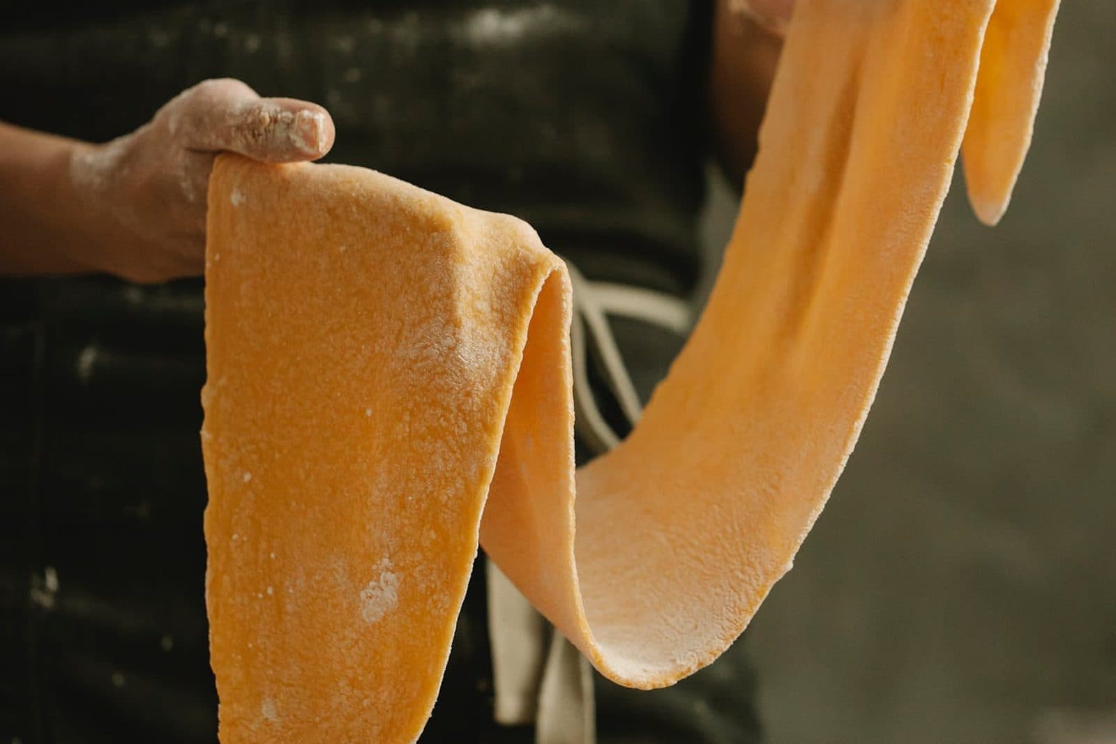 Fresh pasta dough sheet being stretched by hand with flour on a gray work surface