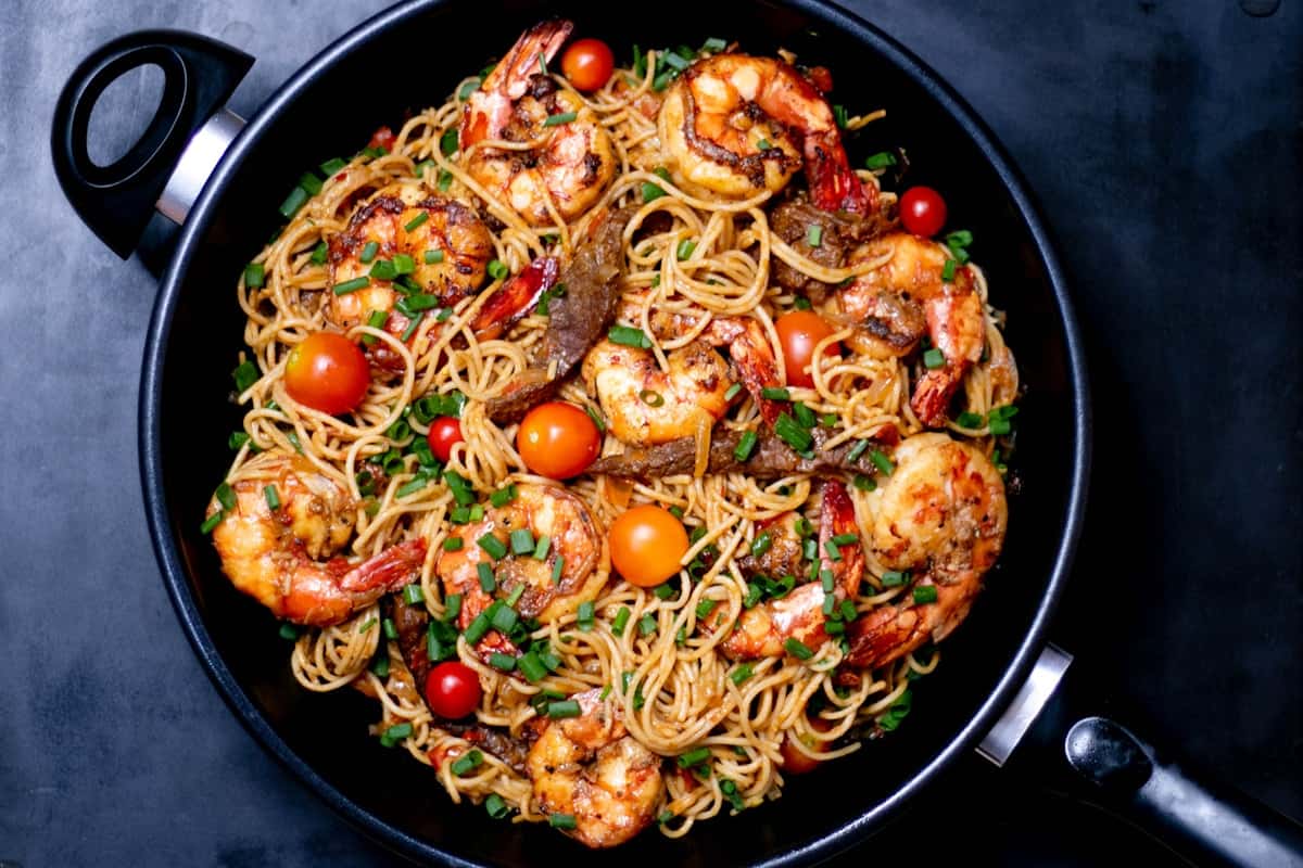 Garlic butter shrimp pasta with noodles, shrimp, tomatoes, and scallions in a dark pan