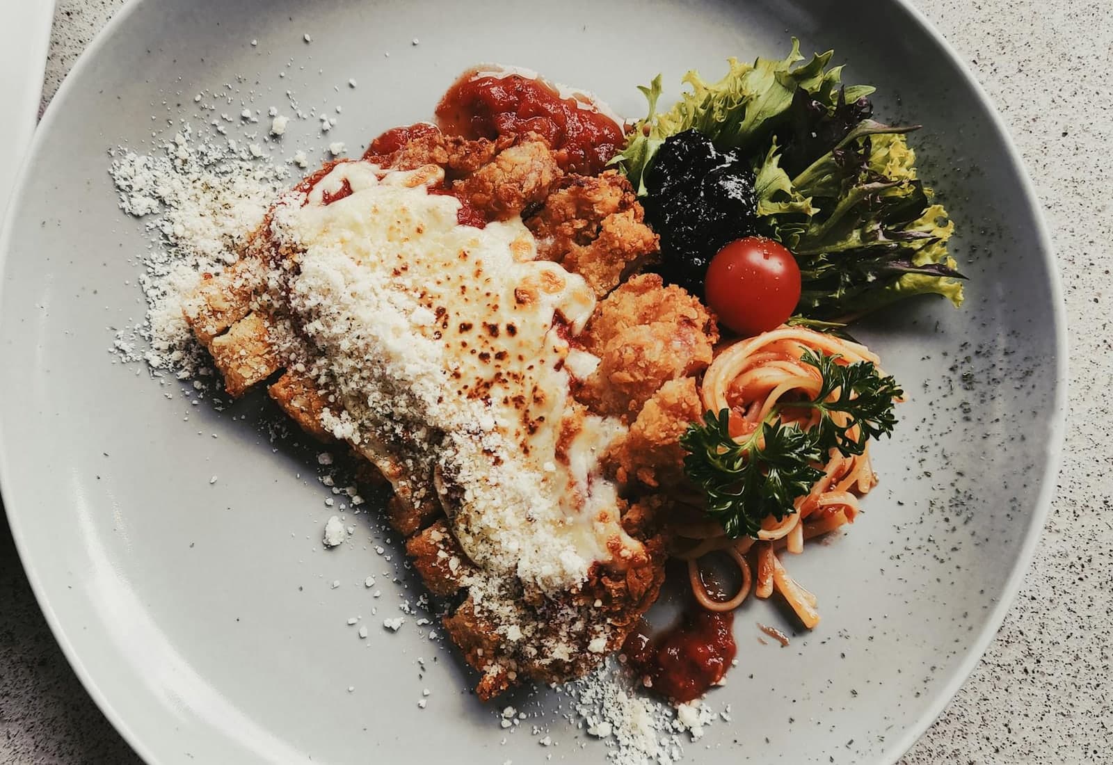 Chicken parmesan-style cutlet with tomato sauce, cheese, pasta, and greens on a grey plate