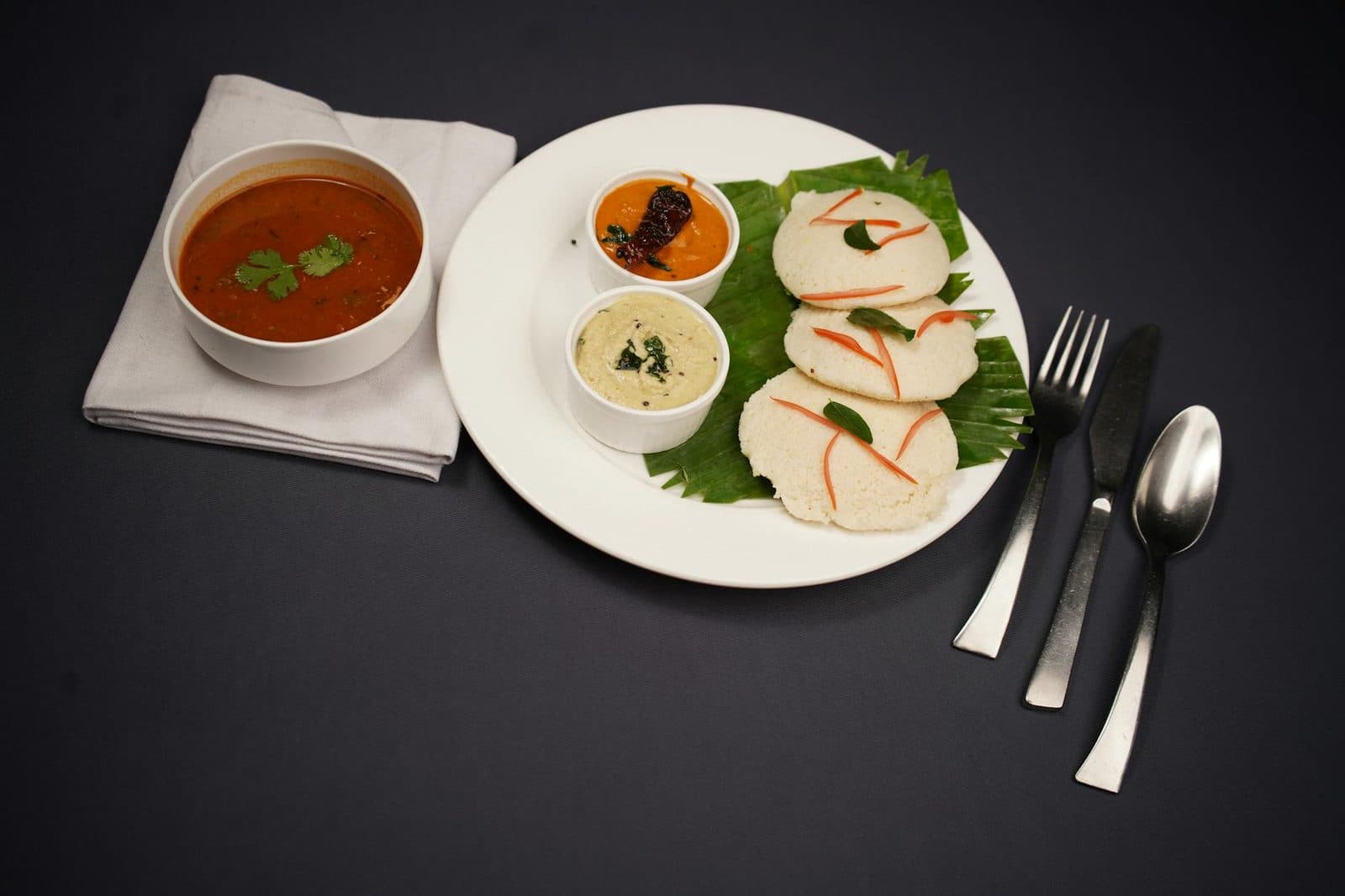 South Indian idli served with sambar and chutneys on a dark plate