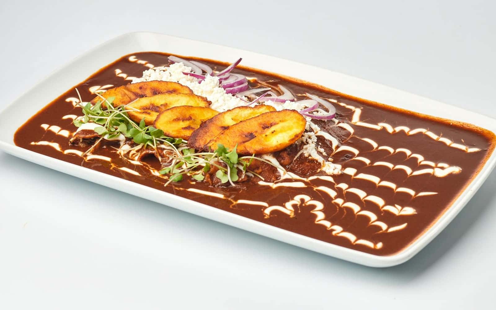 Mole sauce spread on a plate with plantains, cheese, herbs, and dark chile sauce