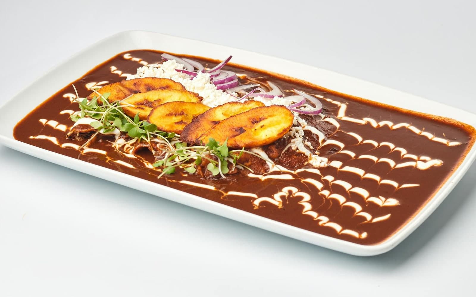 Mole sauce spread on a plate with plantains, cheese, herbs, and dark chile sauce