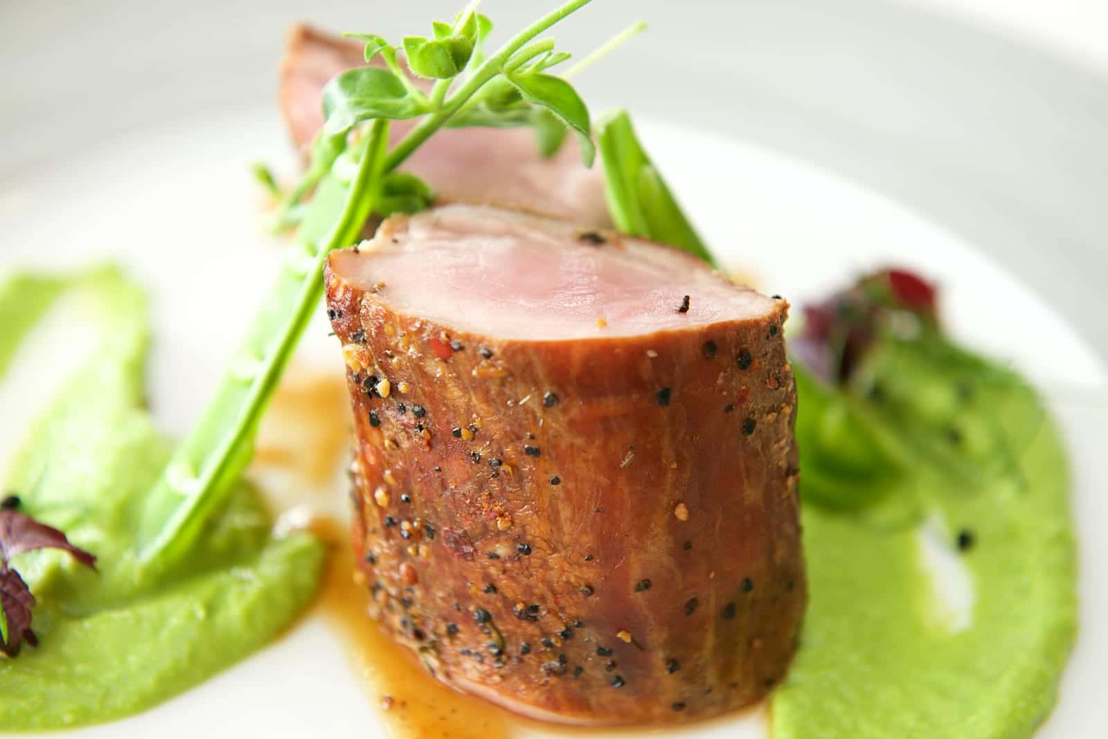 Sliced herb-crusted pork tenderloin with green puree and a small salad on a white plate