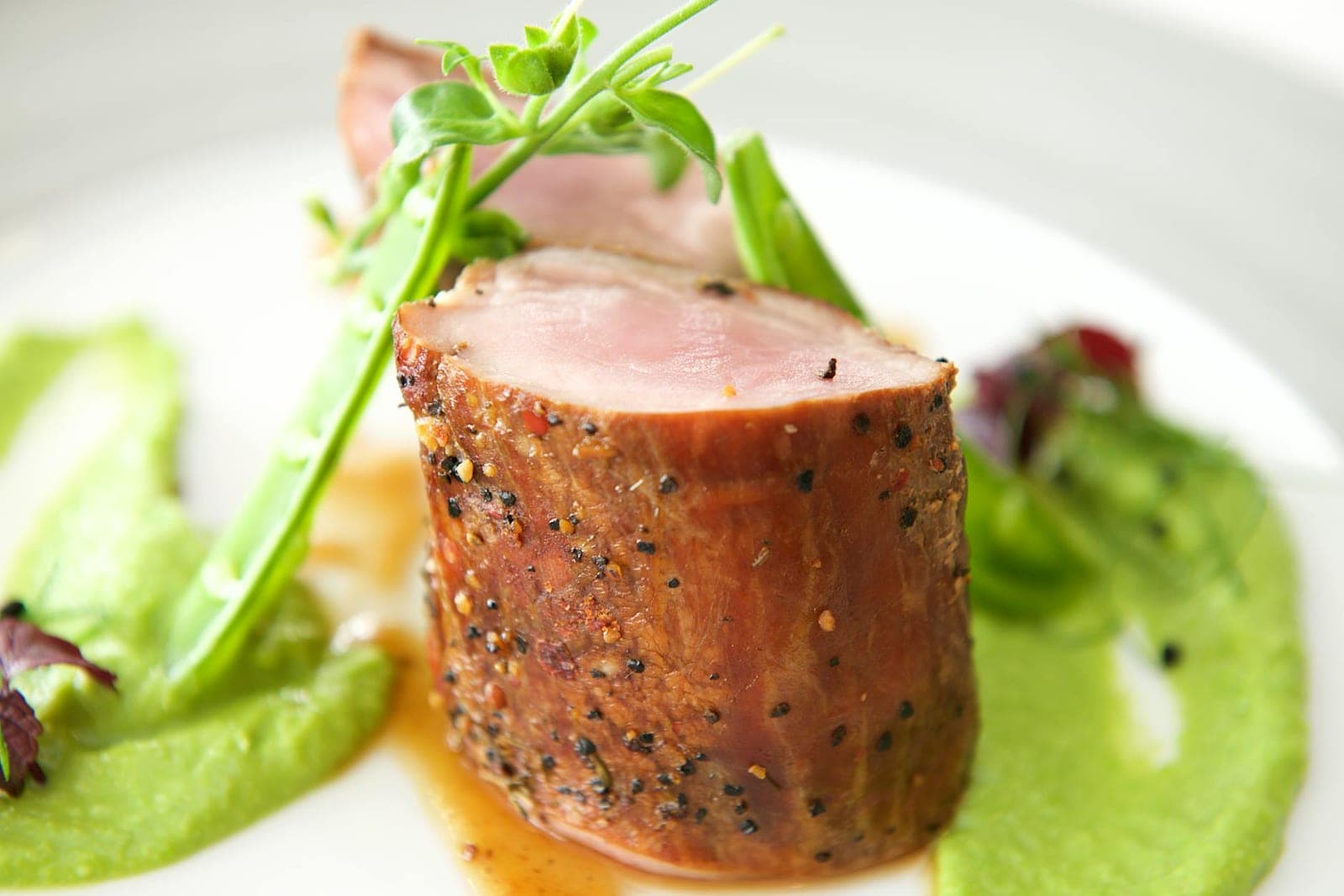 Sliced herb-crusted pork tenderloin with green puree and a small salad on a white plate