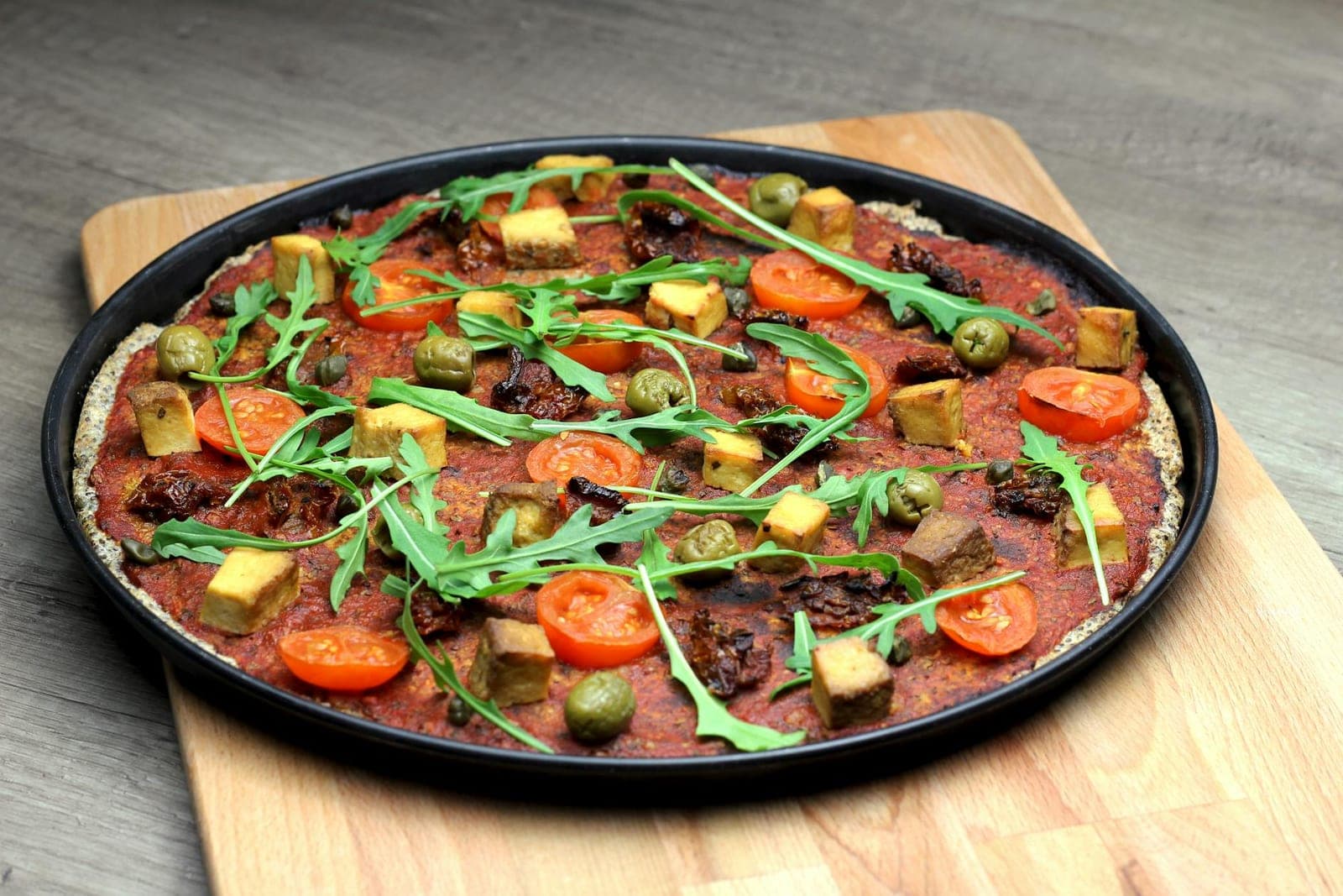 Vegan pizza topped with tomatoes, greens, olives, tofu, and vegetables on a dark pan