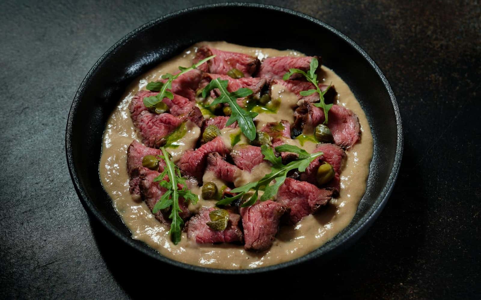 Vitello tonnato with sliced veal, tuna sauce, arugula, and herbs in a dark bowl