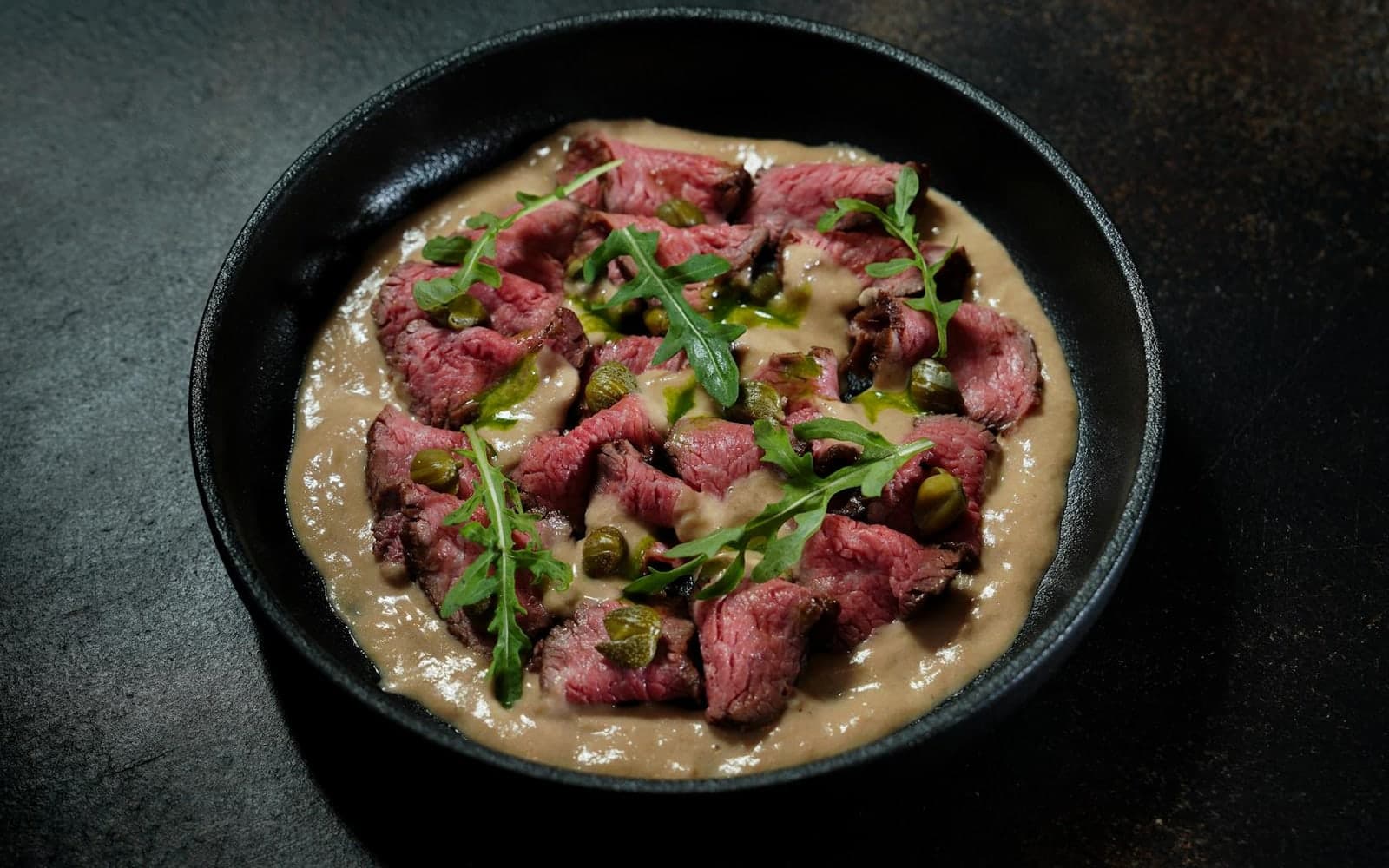 Vitello tonnato with sliced veal, tuna sauce, arugula, and herbs in a dark bowl